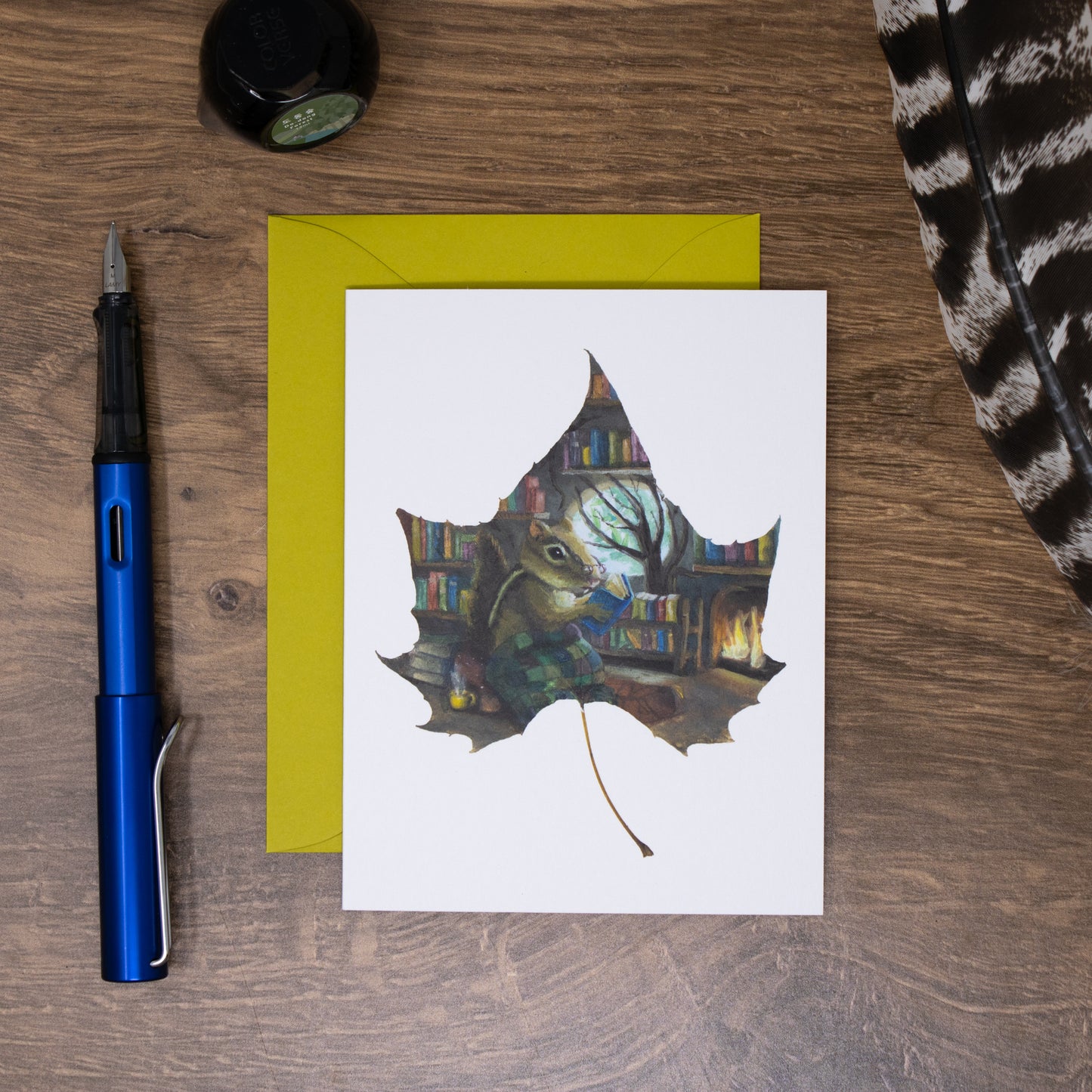 Greeting card with a chipmunk on a leaf design, pen, and envelope on a wooden surface