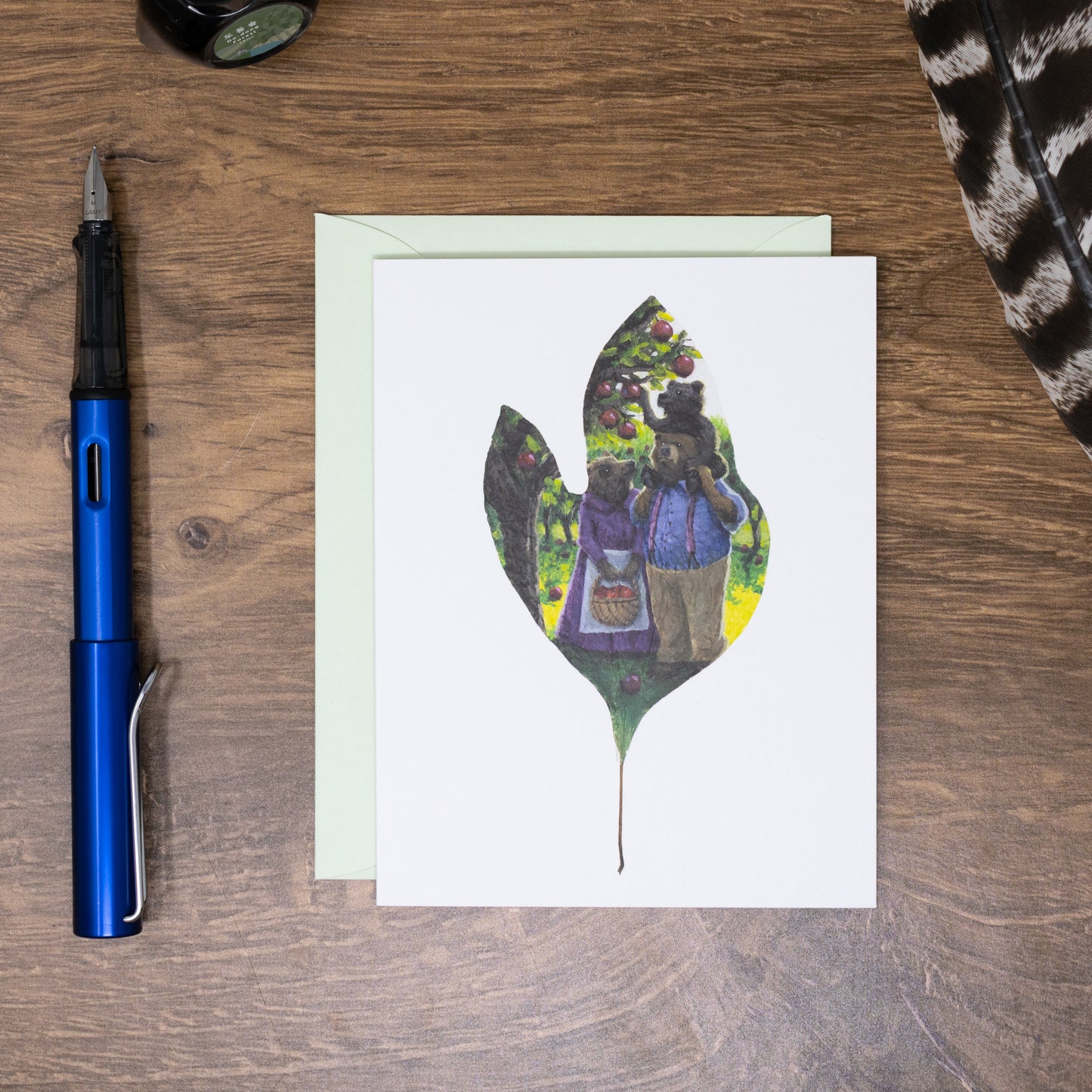 Greeting card with leaf design featuring a bear family, placed on a wooden surface with a pen and other items.