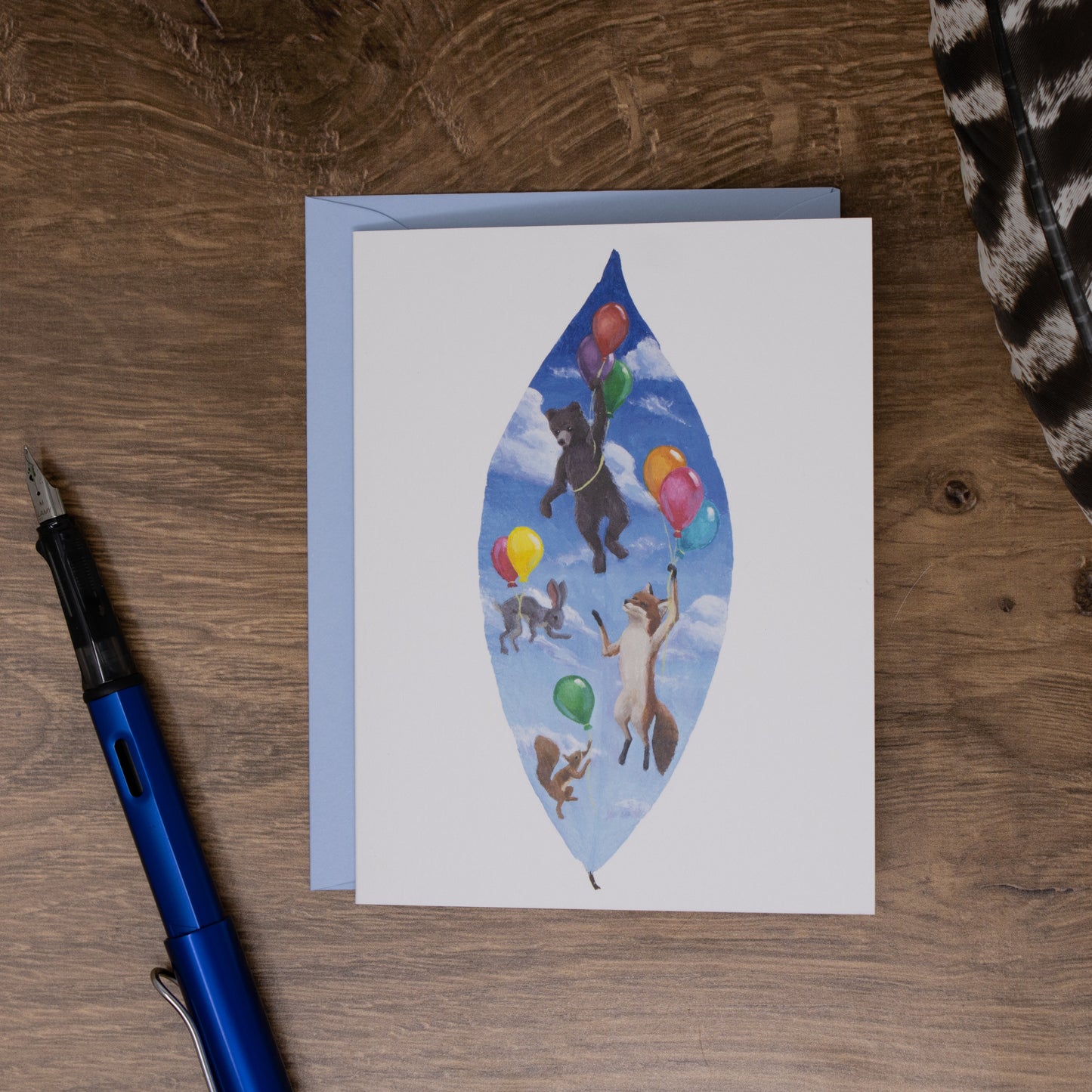 Greeting card with animal illustration of bears and balloons on a wooden surface with a pen.