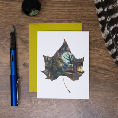 Greeting card with a chipmunk on a leaf design, pen, and envelope on a wooden surface