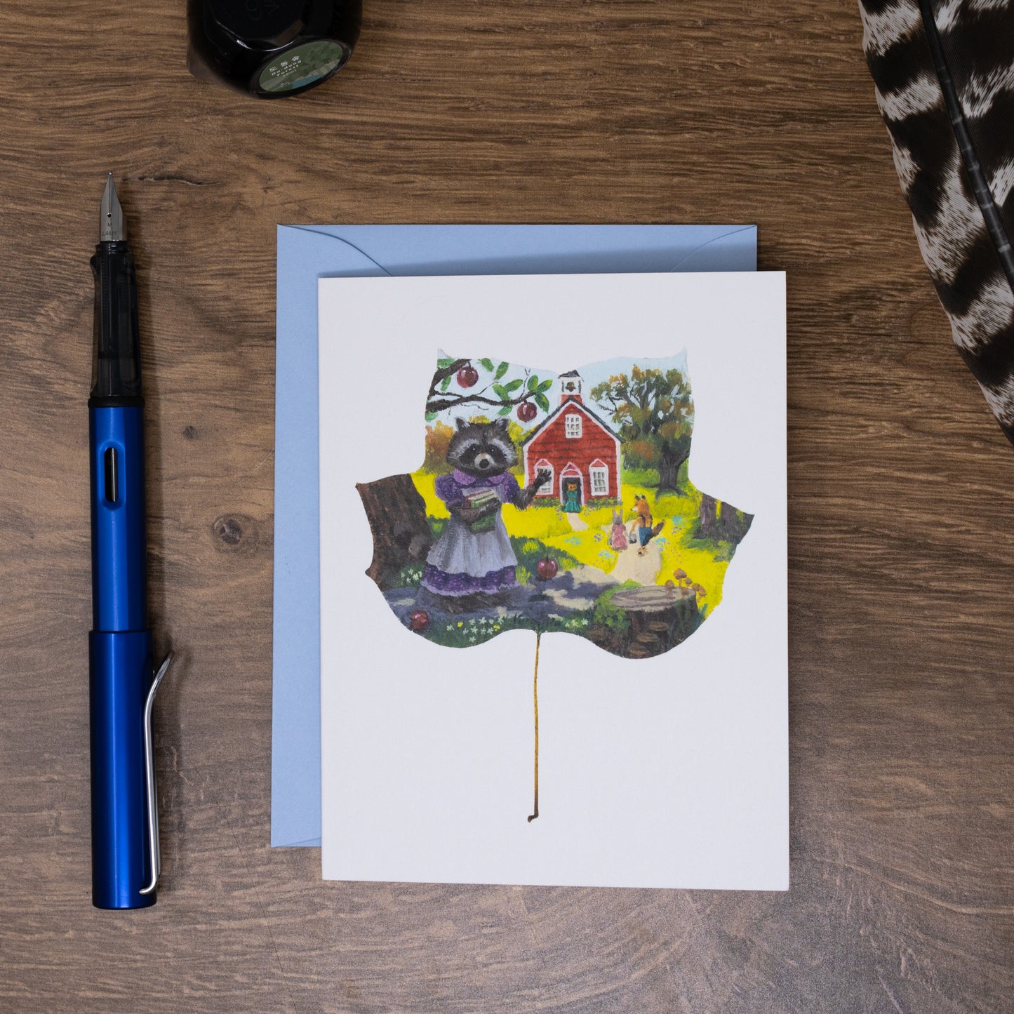 Greeting card with a raccoon on a wooden surface with a pen and envelope.