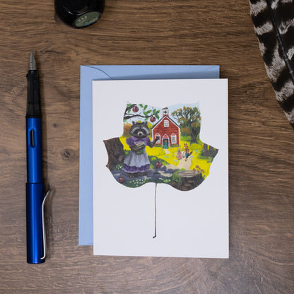 Greeting card with a raccoon on a wooden surface with a pen and envelope.
