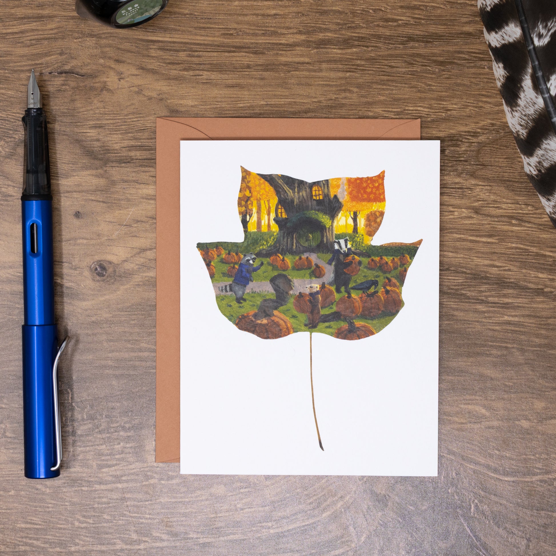 Greeting card with autumn-themed illustration of a leaf on a wooden surface with a pen and cat.