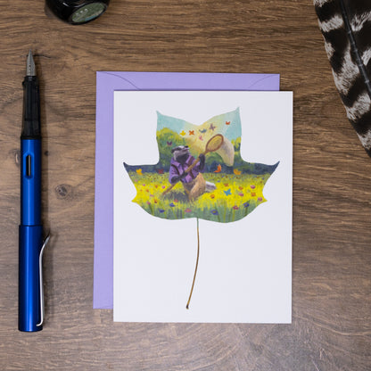 Greeting card with a leaf design featuring a badger and butterfly, placed on a wooden surface with a pen and fabric.