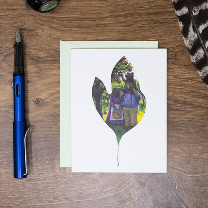Greeting card with leaf design featuring a bear family, placed on a wooden surface with a pen and other items.