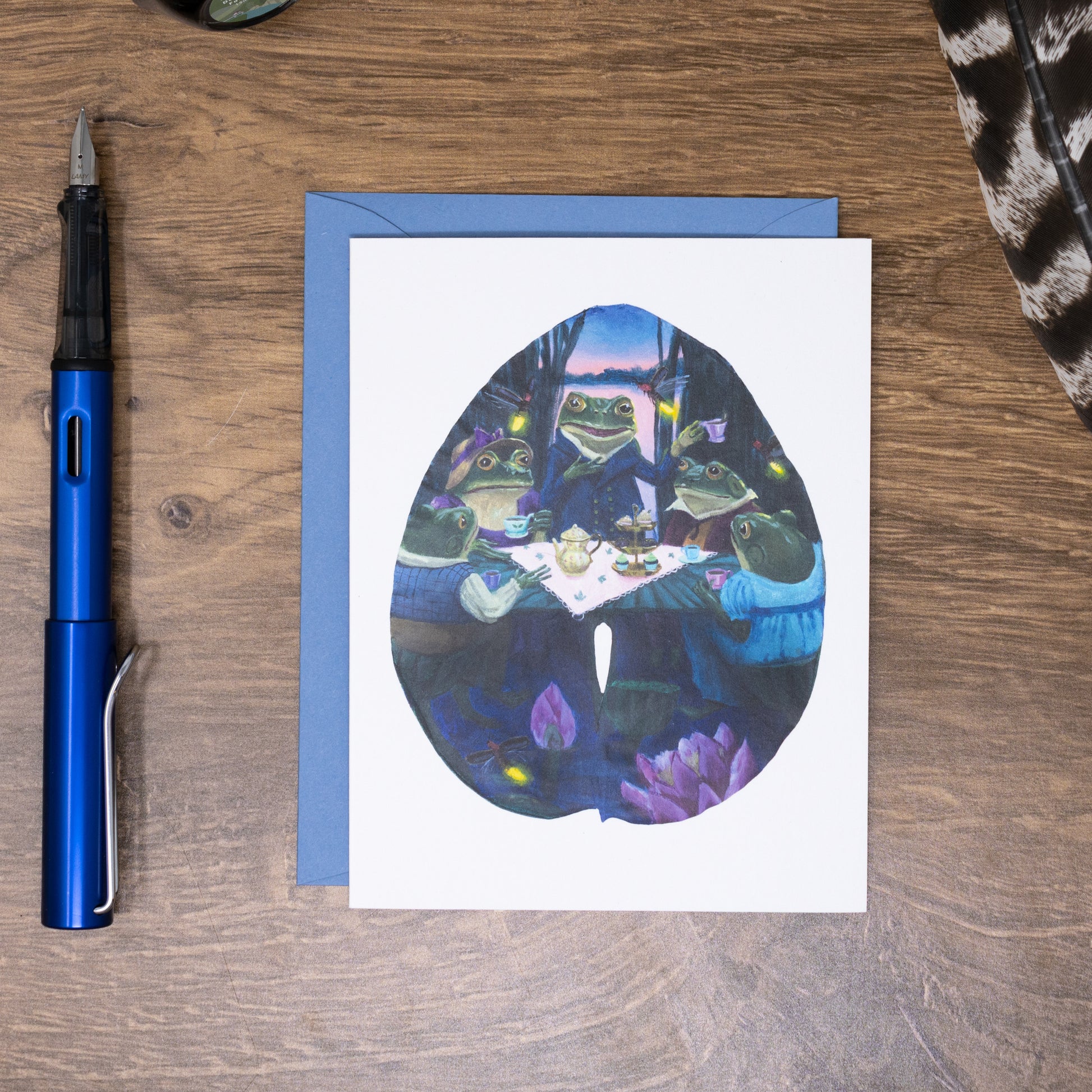 Greeting card with frog illustration on a wooden surface with a pen and envelope.